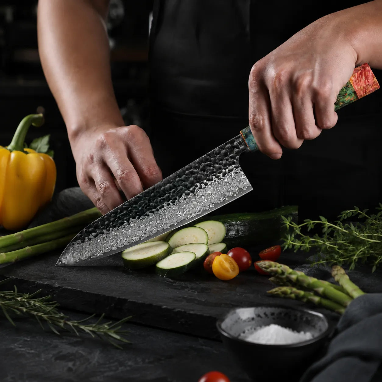 Person using a chef's knife to slice vegetables on a dark surface with various ingredients around.